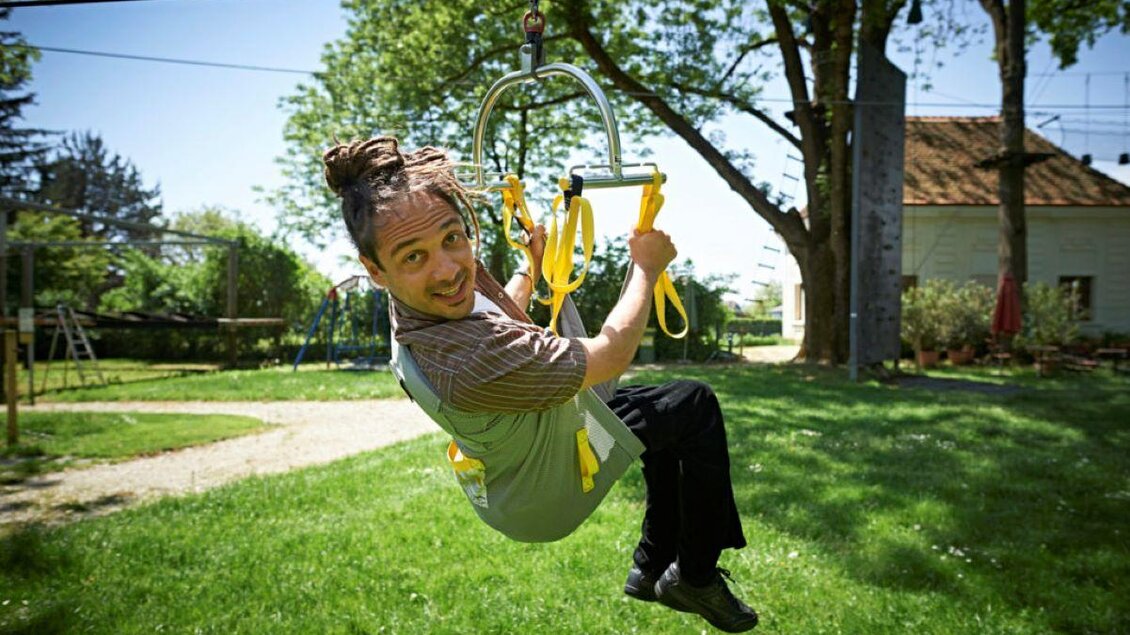 Ein fröhlicher Mann schwingt an einer Zipline im Freien. Die grüne Wiese und Bäume im Hintergrund verleihen der Szene eine lebendige Atmosphäre. | © Jeseničnik Tomo