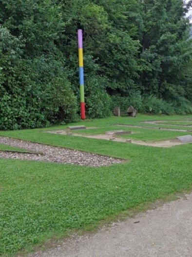 Ein grüner Park mit Steinen in Form von Buchstaben. Im Hintergrund stehen Bäume und ein bunter Mast. | © Kneippanlage Maria Rastl