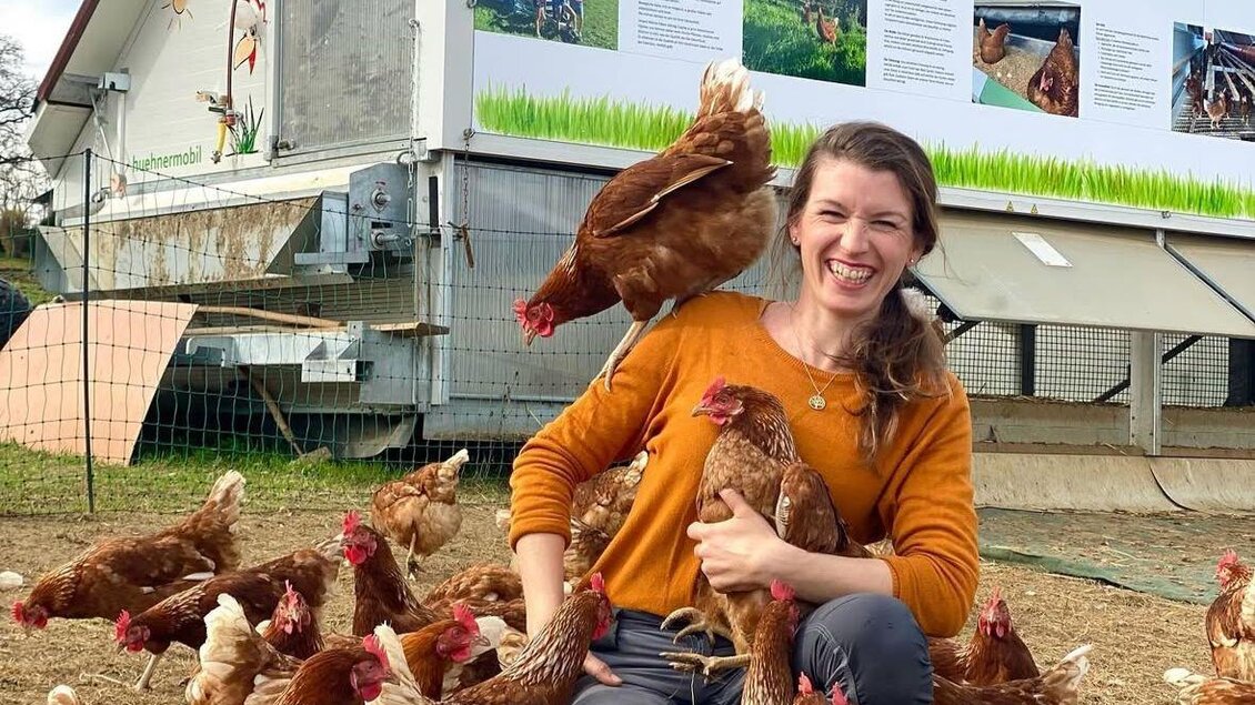 Eine Frau sitzt inmitten von Hühnern und lächelt. Im Hintergrund ist ein Gebäude mit einem bunten Wandbild zu sehen. | © Knippihof