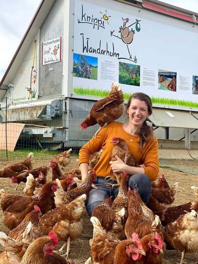 A woman is sitting among chickens and smiling. In the background, there is a building with a colorful mural. | © Knippihof