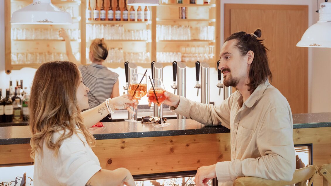 Ein Paar stößt mit Getränken an einer Bar an. Im Hintergrund ist eine Kellnerin zu sehen. | © www.zloam.at