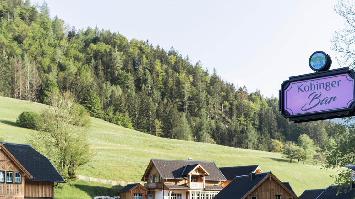 Eine malerische Landschaft mit grünen Hügeln und traditionellen Holzhäusern. Im Vordergrund ist ein Schild mit der Aufschrift "Kobinger Bar" zu sehen. | © www.zloam.at