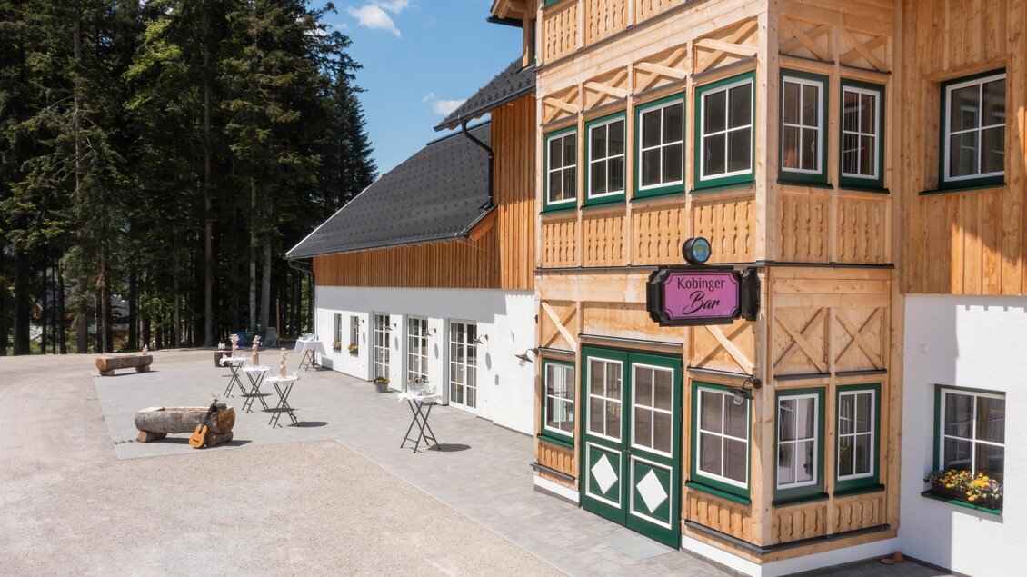 Ein modernes Gebäude mit holzverkleideter Fassade in einer Waldumgebung. Vor dem Gebäude stehen einige Tische und Stühle auf einem gepflasterten Bereich. | © www.zloam.at