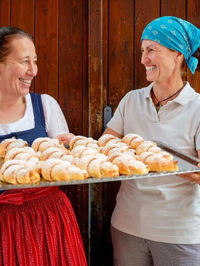 Two women are smiling at each other and holding a tray of fresh croissants. The background is a rustic wooden wall. | © Susanne Enzenberger Red Bull Mediahouse