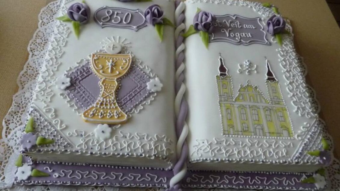Eine kunstvolle Torte in Form eines aufgeschlagenen Buches, dekoriert mit lila Rosen und einem goldenen Kelch. Das Design feiert ein besonderes Jubiläum mit der Aufschrift "350". | © Helmut Kaiser