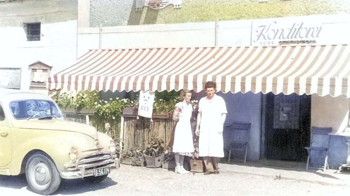 Ein altes Café mit einem gelben Auto davor. Zwei Personen in weißen Schürzen stehen vor dem Eingang. | © Helmut Kaiser
