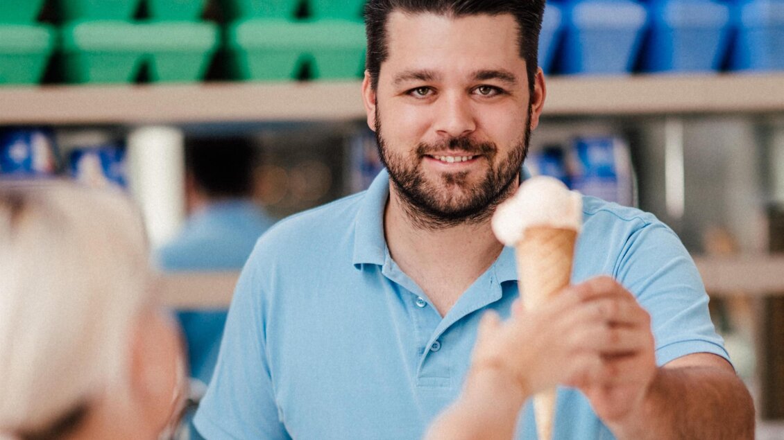 Ein freundlicher Eisverkäufer reicht eine Eistüte an eine Kundin. Im Hintergrund sind verschiedene Eiscremesorten zu sehen. | © Koppitzeis