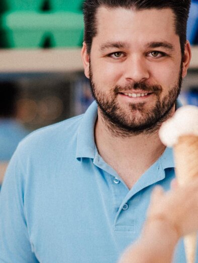 A friendly ice cream vendor hands an ice cream cone to a customer. In the background, various flavors of ice cream are visible. | © Koppitzeis