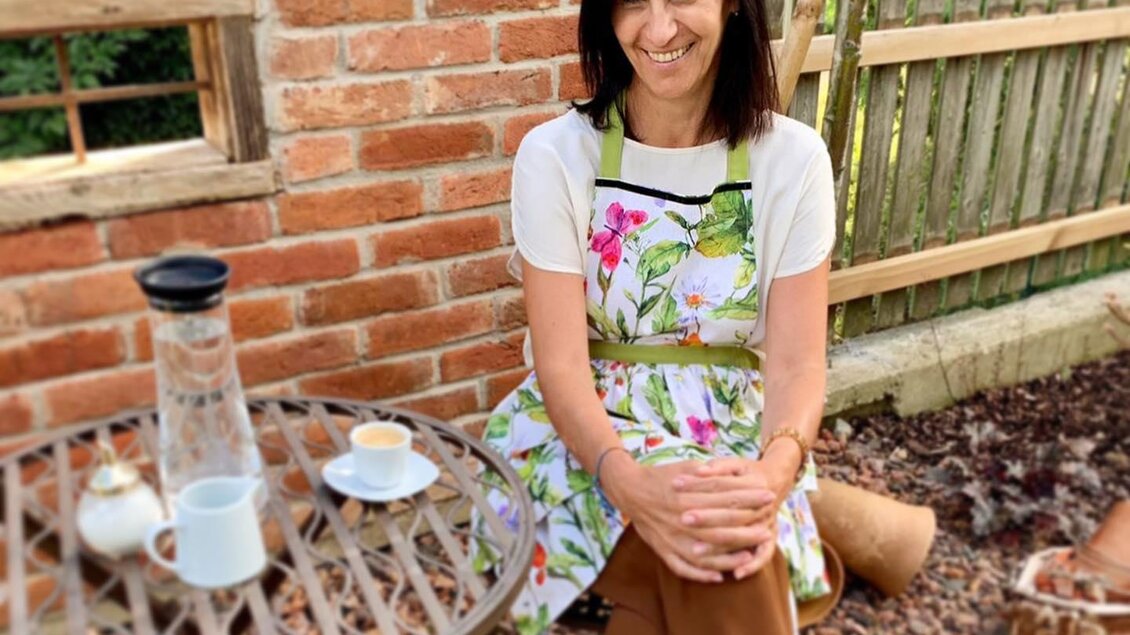 Eine Frau sitzt an einem Tisch im Freien, bekleidet mit einer blumigen Schürze. Auf dem Tisch stehen Kaffeetassen und eine kleine Kaffeekanne. | © KostBar Café & Greißlerei