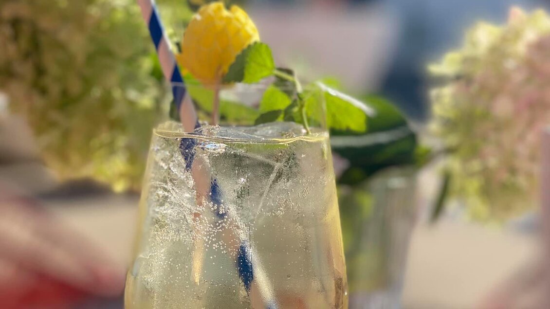 Ein erfrischender Cocktail in einem Glas mit einem bunten Strohhalm. Im Hintergrund blühen grüne und rosa Blumen. | © KostBar Café & Greißlerei