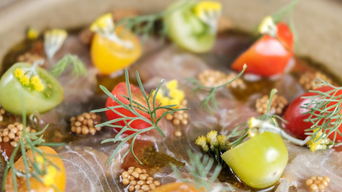 Ein kunstvoll angerichteter Teller mit dünn geschnittenem Fisch, garniert mit bunten Kirschtomaten und Senfsamen. Im Hintergrund ist eine kleine Schüssel mit Dipsaucen zu sehen. | © Kostbar, Thomas Gartner