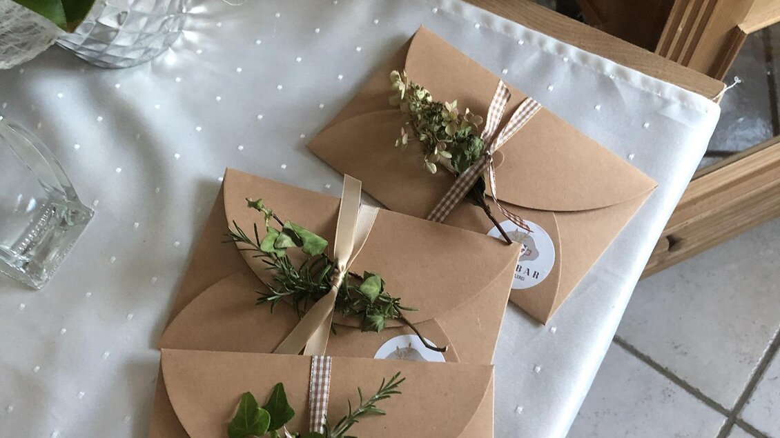Drei elegante Umschläge aus braunem Kraftpapier liegen auf einem weißen Tisch. Jeder Umschlag ist mit einem Band und frischen Kräutern dekoriert. | © KostBar Café & Greißlerei