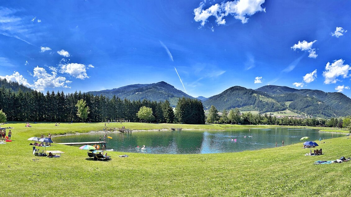 Ein malerischer See umgeben von grünen Wiesen und Bäumen. Im Hintergrund sind majestätische Berge und ein klarer blauer Himmel sichtbar. | © Markus Oberländer