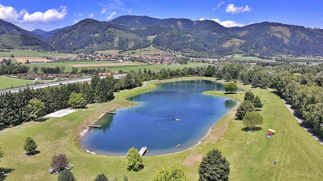 Ein schöner See umgeben von grünen Wiesen und Bäumen. Im Hintergrund erheben sich sanfte Berge unter einem klaren blauen Himmel. | © Markus Oberländer