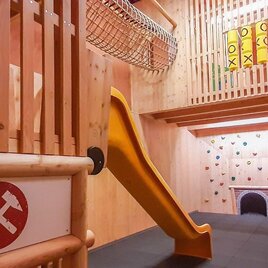 Ein Spielbereich mit einer Rutsche, Kletterwand und einem Netz zum Spielen. Die Wände sind aus Holz und es gibt bunten Klettersteine. | © JUFA Hotels