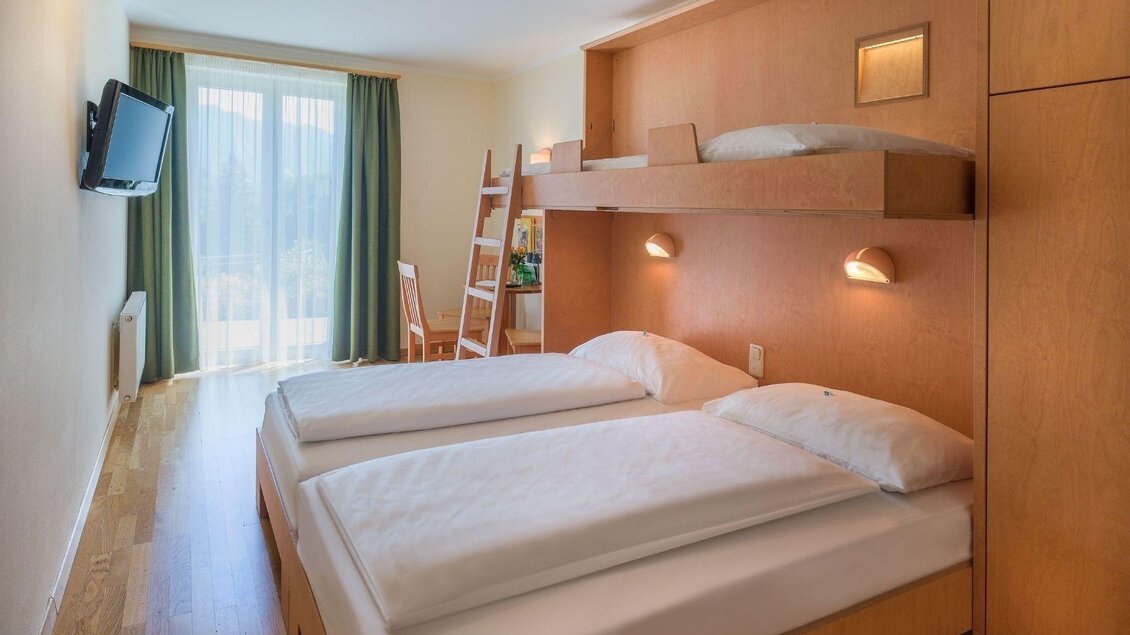 Ein helles Zimmer mit zwei Einzelbetten und einem oberen Schlafbereich. Große Fenster sorgen für viel Tageslicht und eine freundliche Atmosphäre. | © JUFA Hotels