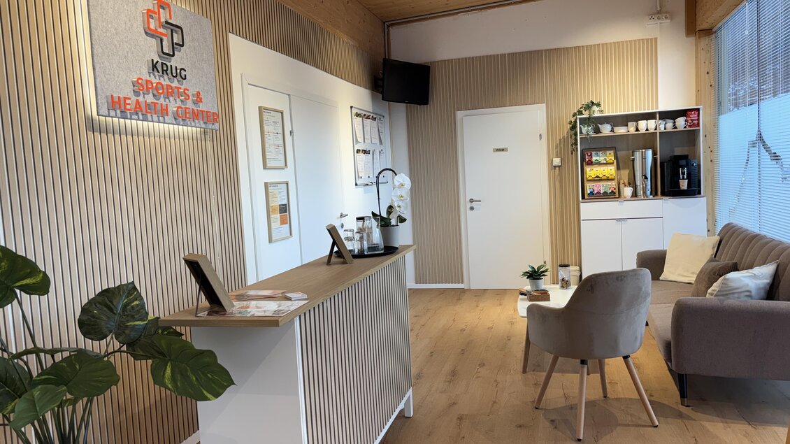 Ein modernes Empfangsbereich mit einem Empfangstresen und bequemen Sitzgelegenheiten. Die Wände sind in hellen Tönen gehalten und es sind einige Pflanzen dekorativ platziert. | © Krug Sports- & Health Center Ramsau