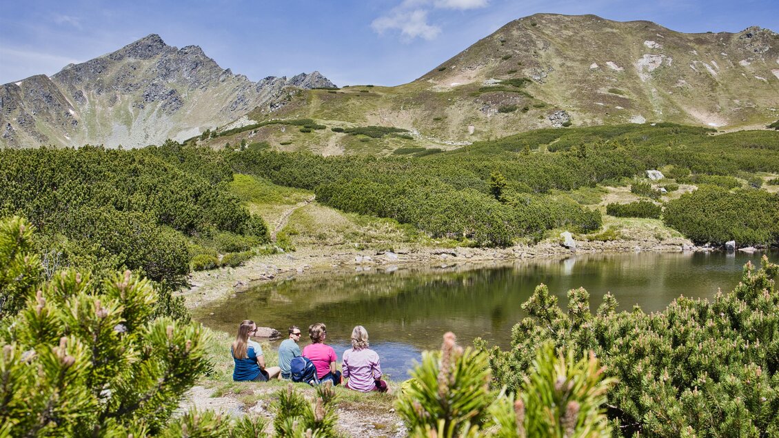 Ein Gruppe von Menschen sitzt am Ufer eines klaren Sees in den Bergen. Die Szene ist umgeben von grünen Wäldern und majestätischen Bergen im Hintergrund. | © Weges OG