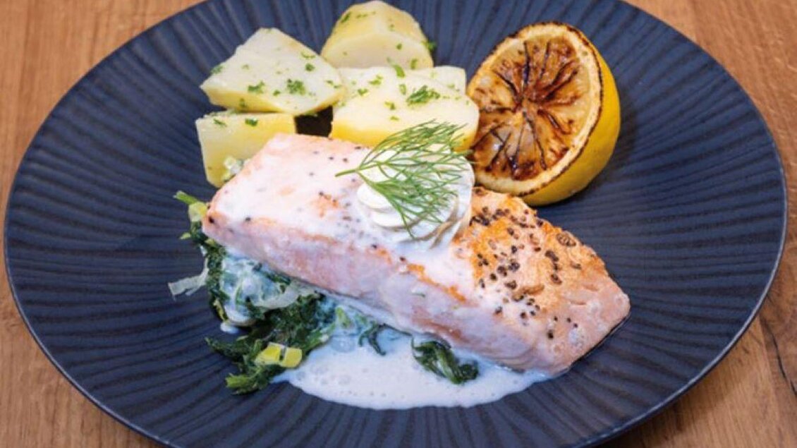 Ein Stück gegrillter Lachs auf einem blauen Teller, serviert mit Kartoffeln und einer gegrillten Zitrone. Dazu gibt es eine cremige Soße und frisches Gemüse. | © Kulinarkwerk Fresh
