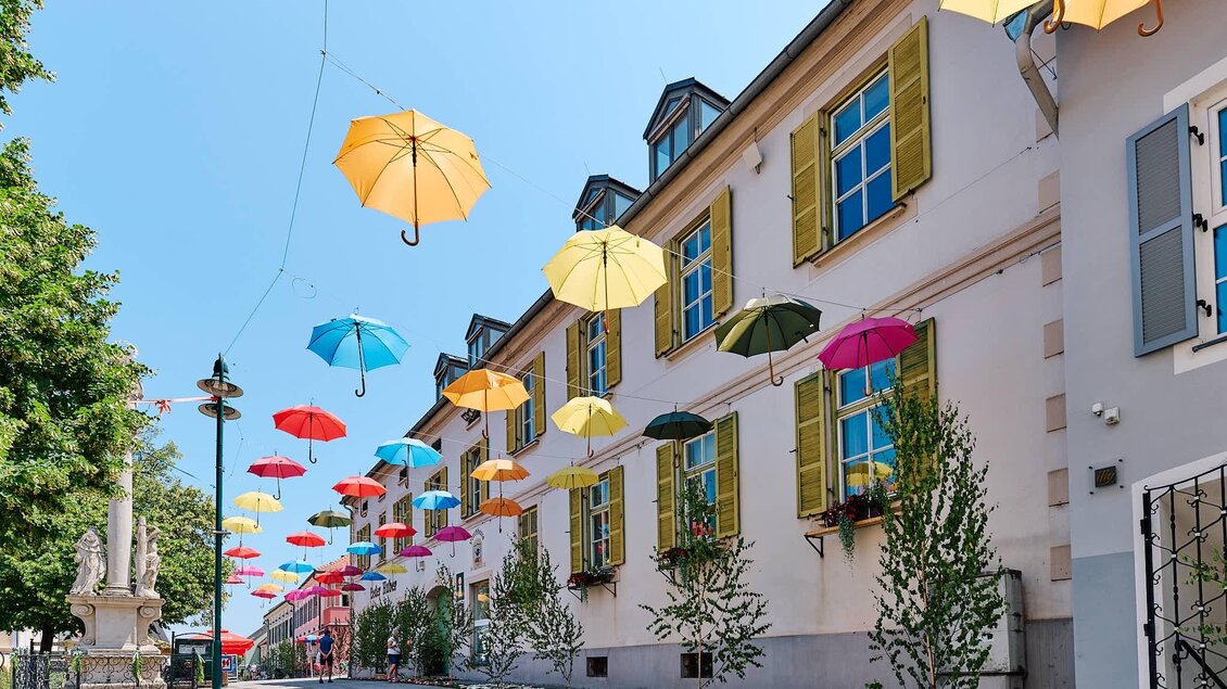 Eine malerische Straße mit bunten Regenschirmen, die über die Häuser hängen. Die Gebäude haben große Fenster und leuchtend gelbe Fensterläden. | © Kultur- & Museumsverein Eibiswald