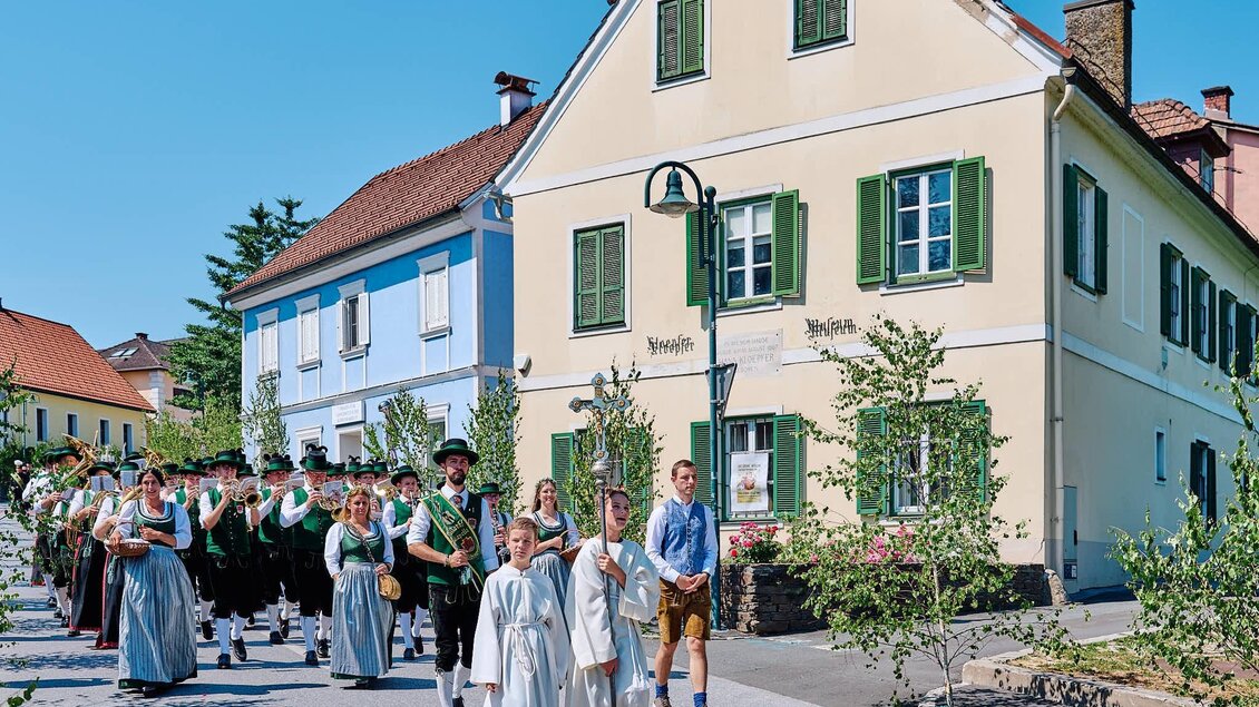 Eine festliche Parade mit Menschen in traditioneller Kleidung zieht durch eine malerische Straße. Im Hintergrund sind bunte Häuser und Bäume zu sehen. | © Kultur- & Museumsverein Eibiswald