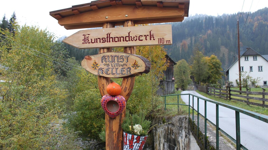 Ein rustikales Holzschild zeigt den Weg zum Kunsthandwerk in 110 m Entfernung. Die Umgebung ist von Bäumen und Häusern umgeben, mit einer ländlichen Straße im Vordergrund.