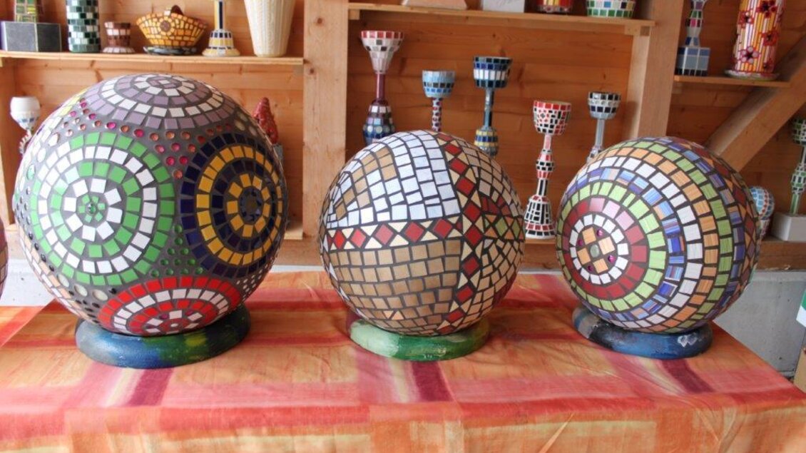 Drei bunte Mosaik-Skulpturen in Form von Kugeln stehen auf einem Tisch. Hintergrund ist eine holzverkleidete Wand mit weiteren dekorativen Gegenständen.