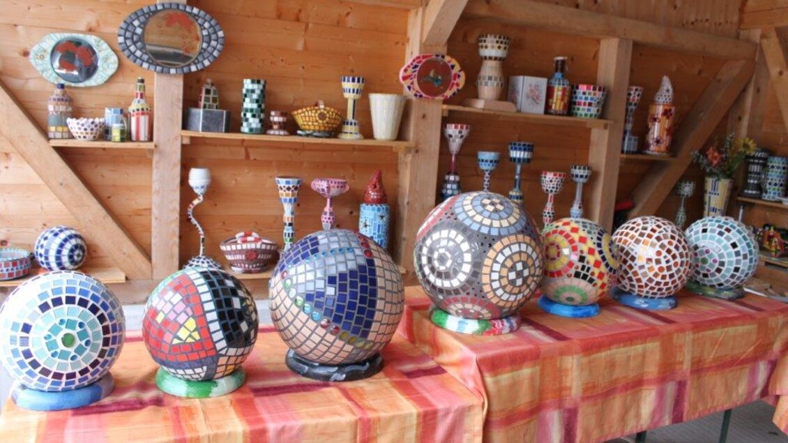 Bunte Mosaikskulpturen in verschiedenen Größen stehen auf einem Tisch. Im Hintergrund sind weitere dekorative Objekte in einem Holzraum zu sehen.