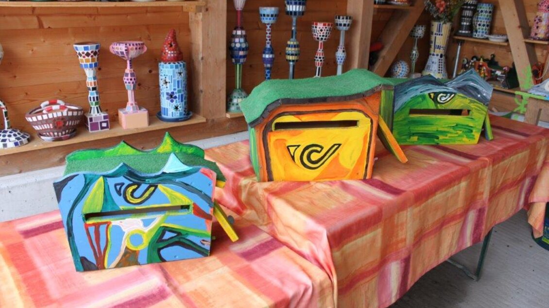 Bunte, handgefertigte Kisten stehen auf einem Tisch. Im Hintergrund sind weitere dekorative Kunstwerke zu sehen.