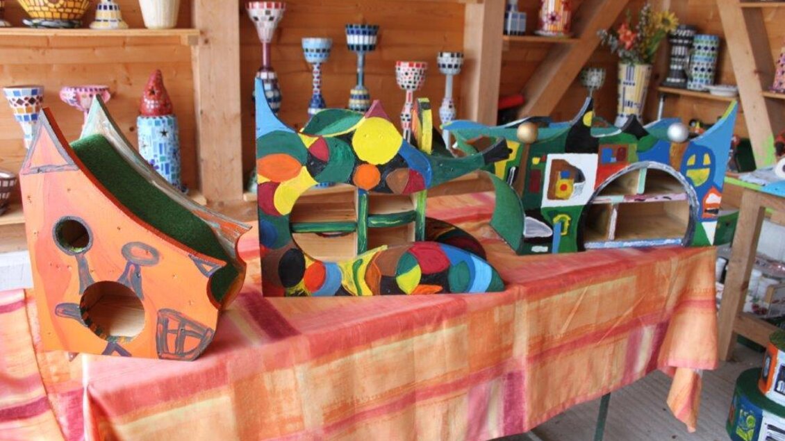 Bunte, handgefertigte Holzskulpturen auf einem Tisch. Im Hintergrund sind weitere dekorative Objekte zu sehen.