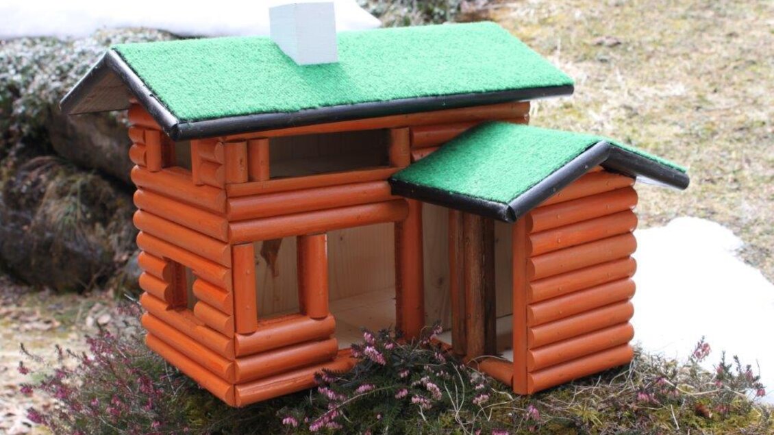 Ein kleines, orangefarbenes Spielhaus im Stil einer Blockhütte mit einem grünen Dach. Es steht in einer Gartenlandschaft mit etwas Schnee im Hintergrund.