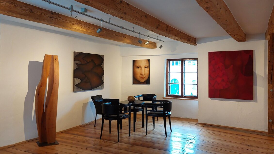 Ein modernes Kunststudio mit Holzfußboden und massiven Holzbalken. An den Wänden hängen verschiedene Gemälde, und in der Mitte steht ein Tisch mit schwarzen Stühlen. | © Romana Leitner