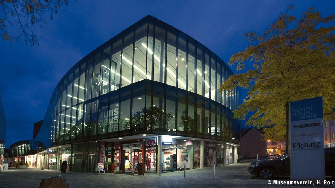 Ein modernes Gebäude mit einer gläsernen Fassade steht bei Nacht im Licht. Umgeben von Bäumen und Geschäften, strahlt es eine einladende Atmosphäre aus. | © Oststeiermark Toruismus