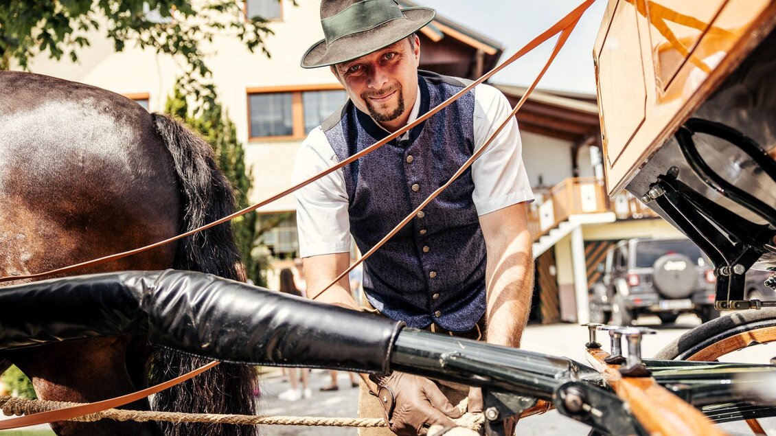 Ein Mann in traditioneller Kleidung arbeitet an einer Pferdekutsche. Im Hintergrund sind ein Haus und Bäume zu sehen. | © Wolfgang Spekner