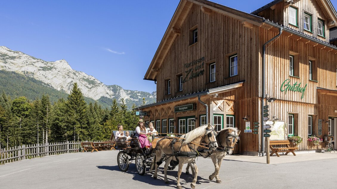 Eine schöne Almhütte aus Holz mit einem Pferdewagen und Passagieren. Im Hintergrund sind Berge und Wälder zu sehen. | © www.zloam.at