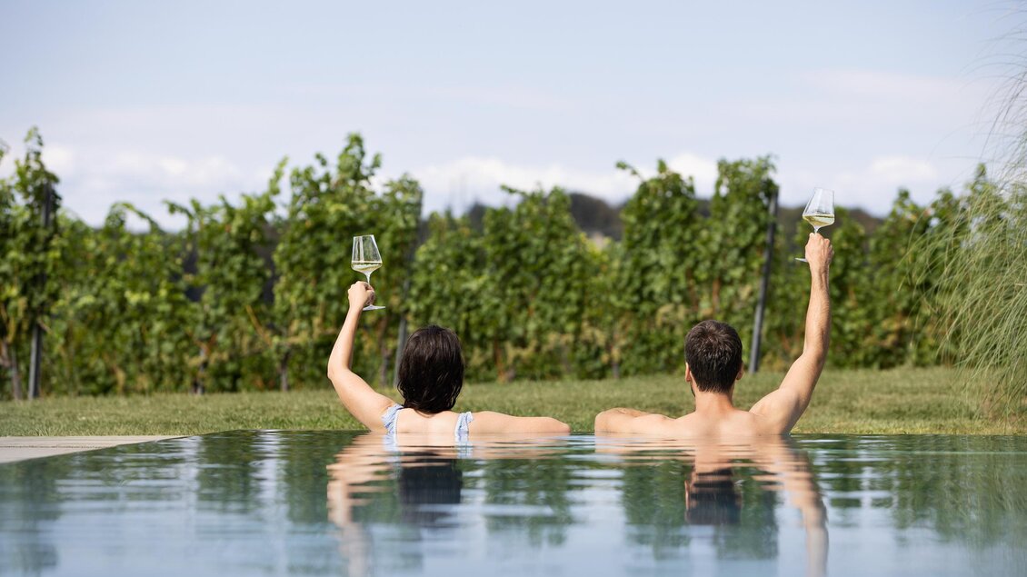 Ein Paar entspannt in einem Pool und hält Glaswein in den Händen. Im Hintergrund sind Weinreben und ein klarer Himmel zu sehen. | © LOISIUM