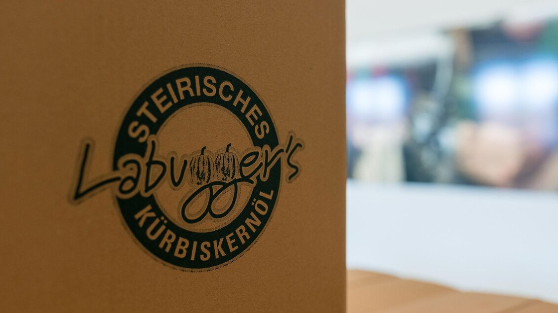 Eine Verpackung für steirisches Kürbiskernöl mit dem Logo von Labugger's. Der Hintergrund zeigt unscharfe Bilder von Produktionsprozessen. | © RM SWS GmbH | webquartier.at