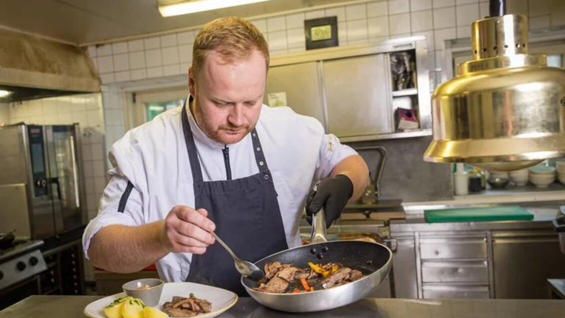 Ein Koch bereitet ein Gericht in einer modernen Küche vor. Auf dem Tisch liegt ein Teller mit Fleisch und Beilagen. | © Wild und Team Fotoagentur GmbH