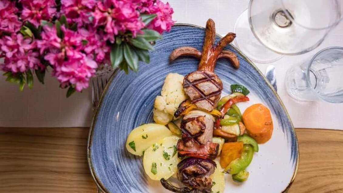 Ein köstliches Gericht aus gegrilltem Fisch, umgeben von buntem Gemüse. Im Hintergrund sind frische Blumen und Gläser zu sehen. | © Wild und Team Fotoagentur GmbH