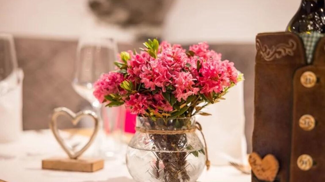 Ein schöner Tisch mit einer Glasvase voller pinken Blumen. Im Hintergrund sind Weingläser und eine Flasche Wein zu sehen. | © Wild und Team Fotoagentur GmbH