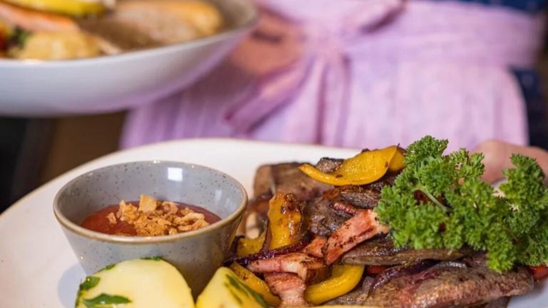 Ein Teller mit gebratenem Fleisch, Kartoffeln und einer würzigen Sauce. Das Gericht wird von einer Person in traditioneller Kleidung serviert. | © Wild und Team Fotoagentur GmbH