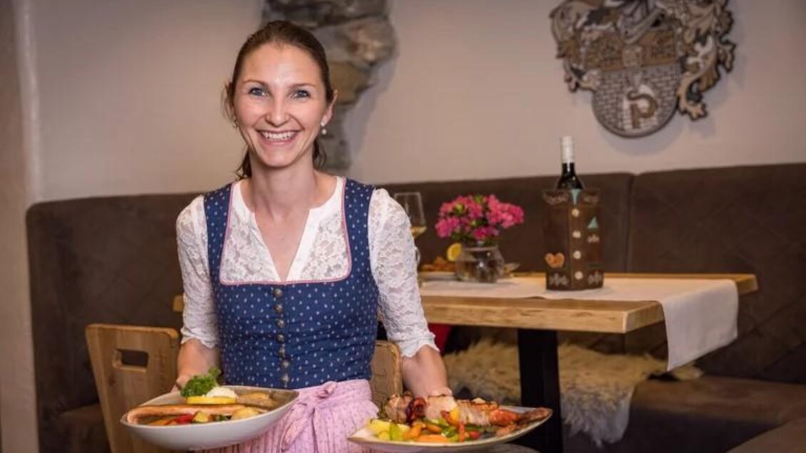 Eine freundliche Kellnerin trägt zwei Teller mit köstlichem Essen. Im Hintergrund ist ein gemütliches Restaurantambiente zu sehen. | © Wild und Team Fotoagentur GmbH