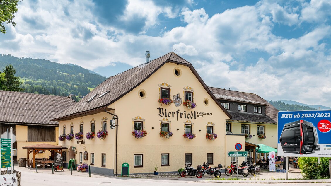 Ein charmantes Gasthaus namens Bierfield umgeben von grünen Bergen und einem klaren Himmel. Die Fassade ist mit Blumen dekoriert und es gibt Bänke im Freien.
