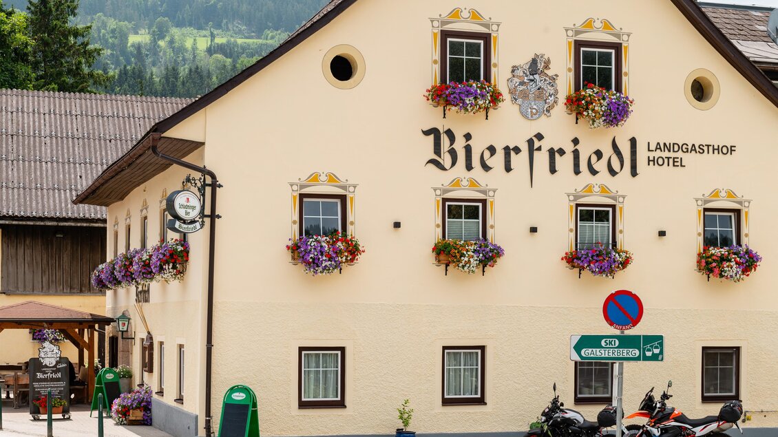 Das Bild zeigt ein charmantes Hotel mit der Aufschrift "Bierfried" und blühenden Fensterkästen. Es befindet sich in einer malerischen Alpenlandschaft.