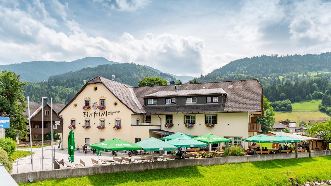 Ein gemütliches Gasthaus mit grünen Sonnenschirmen am Fluss. Im Hintergrund sind sanfte Hügel und ein bewölkter Himmel zu sehen.