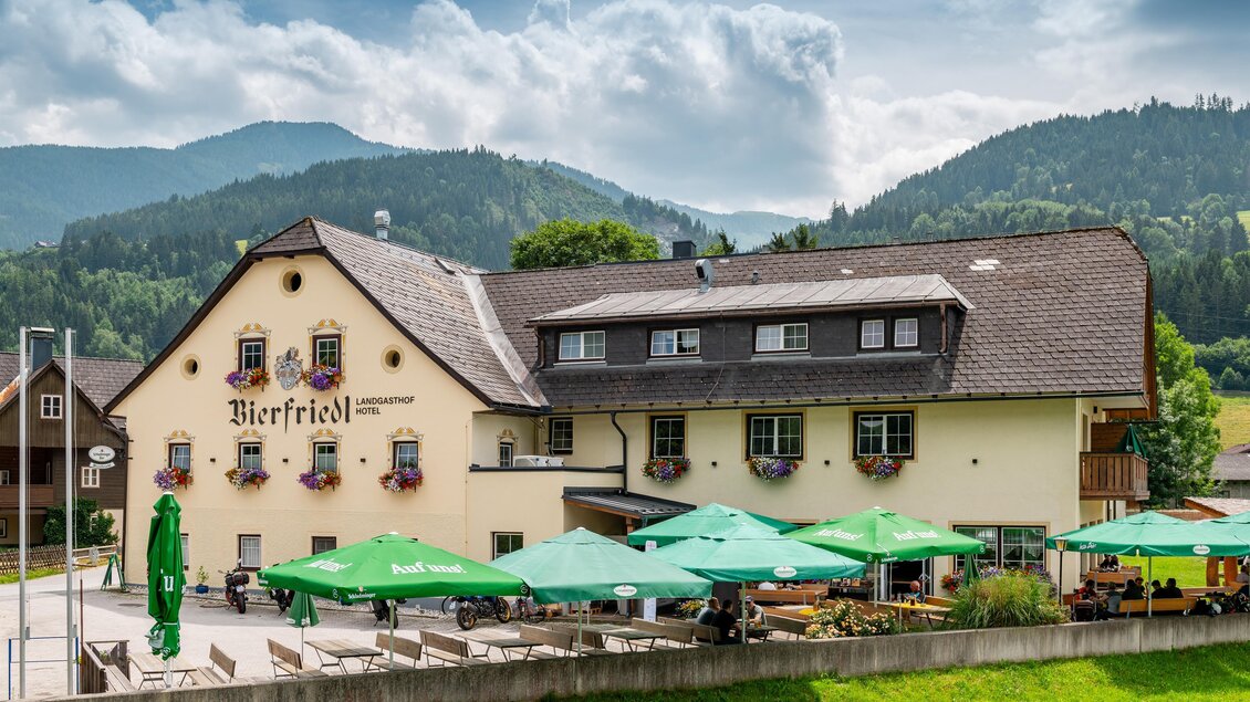 Ein gemütliches Gasthaus namens Bierfriedl mit grünen Schirmen im Freien. Im Hintergrund erstrecken sich sanfte Hügel und eine bewölkte Himmel.