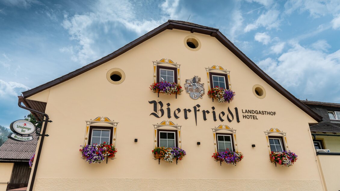 Ein großes, hellgelbes Hotelgebäude mit dem Namen "Bierfriedl" an der Fassade. Blühende Blumenkästen schmücken die Fenster unter einem bewölkten Himmel.