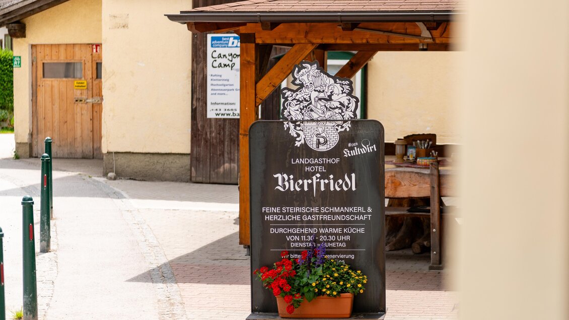 Ein Schild des Hotels Bierfriedl mit einer einladenden Beschreibung der Gaumenfreuden. Im Hintergrund sind traditionelle Gebäude und Blumen sichtbar.