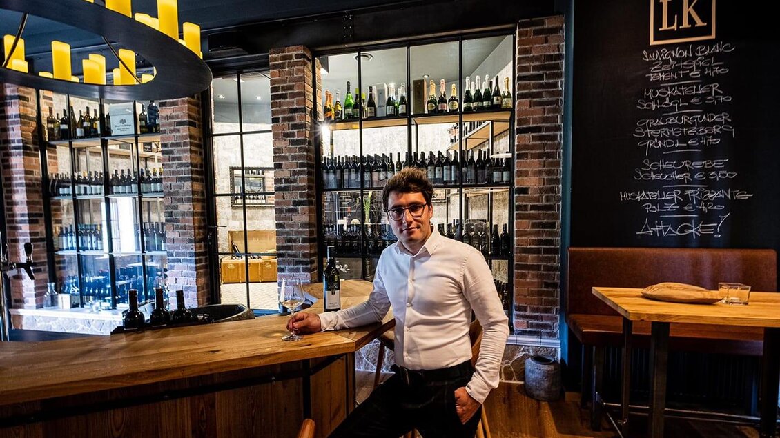 Ein elegantes Restaurant mit einer Holzbar und Weinregalen im Hintergrund. Ein Mann in einem weißen Hemd steht lässig an der Bar. | © Landhaus Kainz | Fam. Kainz