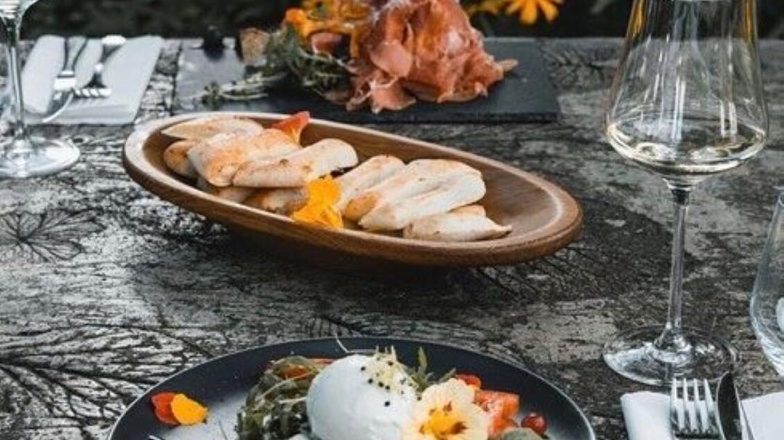 Ein hübsch gedeckter Tisch im Freien mit verschiedenen Gerichten. Im Vordergrund befindet sich ein bunter Salat mit Burrata, im Hintergrund eine Platte mit Schinken und Brot. | © Landhaus Kainz | Fam. Kainz